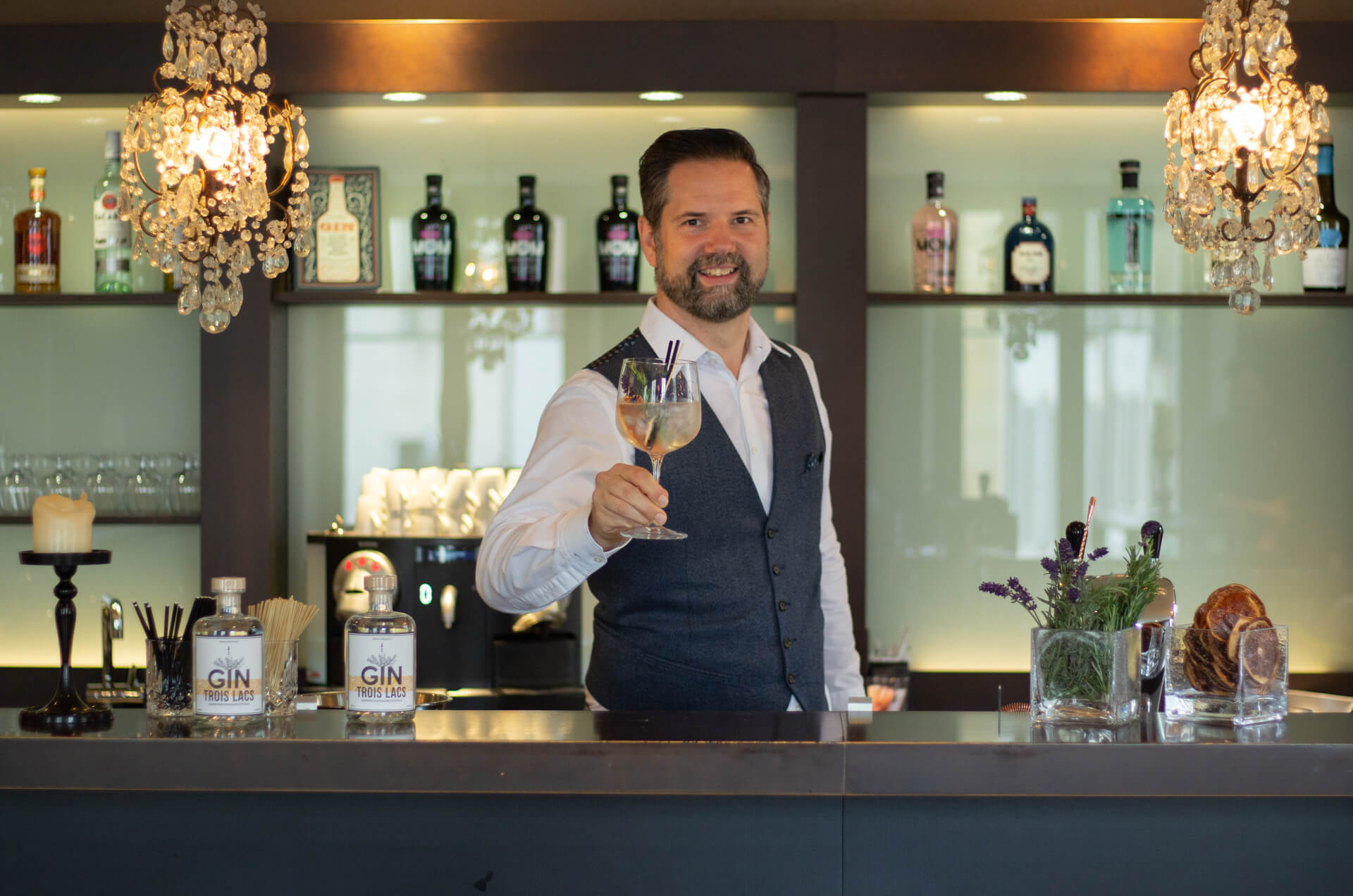 Mann hinter der Bar prostet mit Glas in der Hand der Kamera zu.